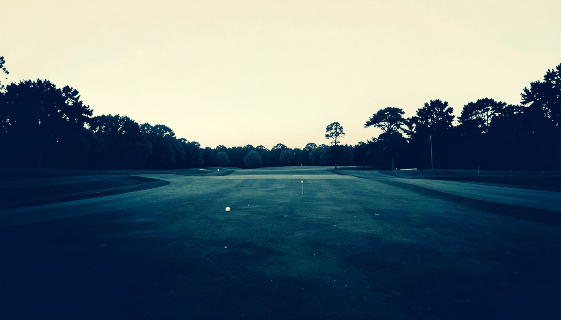 Two-tone duotone photograph of a golf fairway stretching toward distant trees at dusk, rendered in deep navy and warm cream tones