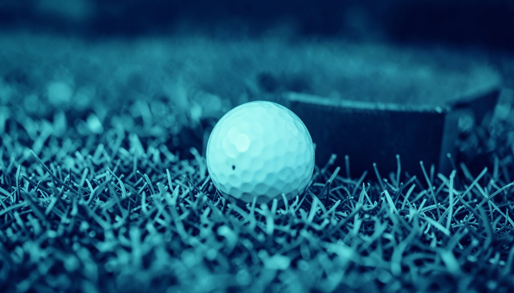 Two-tone duotone photograph of a close-up golf ball resting on short-cut green grass near a dark cup, rendered in deep navy and soft teal tones